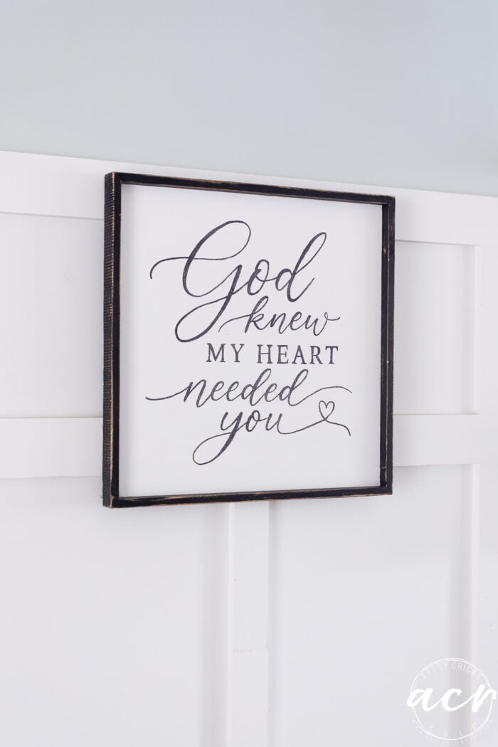 A thrifted redo brings new life to this framed sign on a white paneled wall, where the elegant handwritten script reads, God knew my heart needed you, with a small heart drawing at the bottom right.