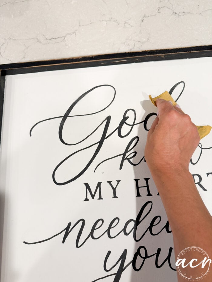 A hand uses sandpaper to smooth the surface of a thrifted sign with black cursive lettering that reads, God Knew My Heart Needed You, giving it a fresh redo.