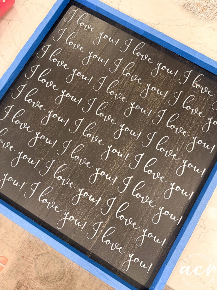 A thrifted redo, this black square sign features “I love you!” repeatedly written in white cursive text, surrounded by blue painters tape along the edges—a heartfelt reminder reminiscent of signs that declare, “God Knew My Heart Needed You.”.