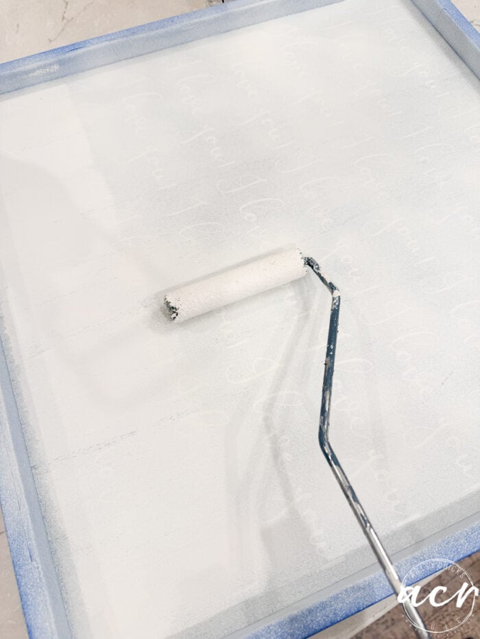 A hand uses a paint roller to apply white paint over a square surface with faint handwritten words visible underneath—perhaps the beginnings of a God Knew My Heart Needed You sign—while blue tape neatly lines the edges, adding a touch of DIY home decor charm perfect for a thrifted redo.