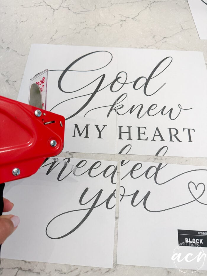 A hand uses a red tape dispenser to join four pieces of paper with elegant script that reads, God Knew My Heart Needed You sign, on a marble surface—perfect for adding sentimental charm to your thrifted decor.