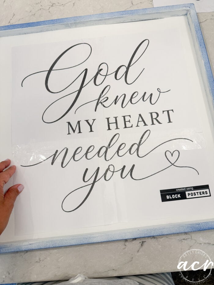 A hand holds a thrifted redo of a paper “God Knew My Heart Needed You” sign, featuring decorative script and a small heart drawing, placed on a white and blue surface.