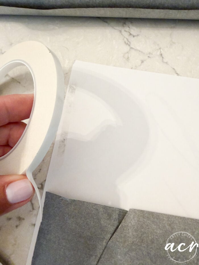 A hand holds double-sided tape next to a white paper with a large cut-out letter, placed on a marble surface—the perfect start for a thrifted redo or a personalized “God Knew My Heart Needed You” sign to elevate your home decor.