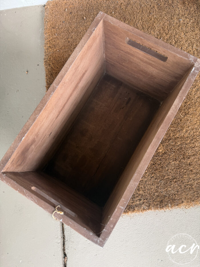 A rectangular wooden harvest crate with handles sits on a textured brown doormat and a concrete floor. The inside of the box is empty, ready to be filled with your favorite fall decor.
