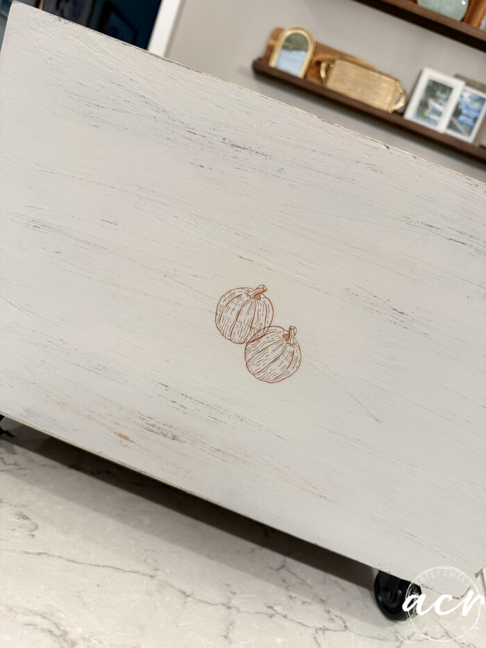 A white wooden surface with a simple line drawing of two pumpkins in the center, creating a cozy Fall Decor accent. The background includes shelves with books and Harvest Crate-inspired decor items.
