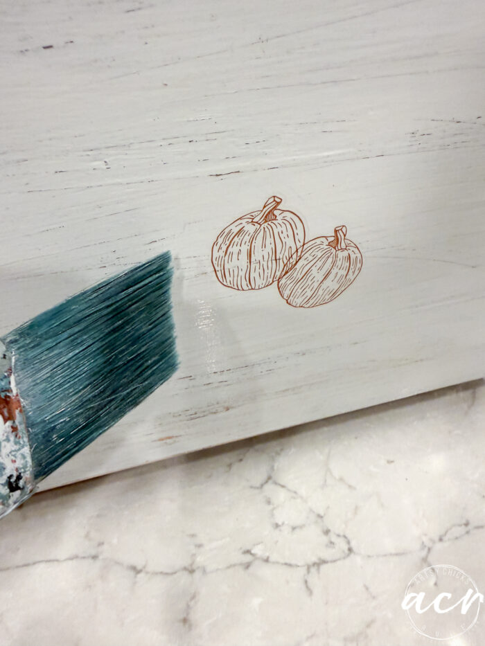 A paintbrush with teal paint is being applied to a white wooden surface with a simple line drawing of two pumpkins, creating charming Fall Decor. The surface rests on a light marble countertop.