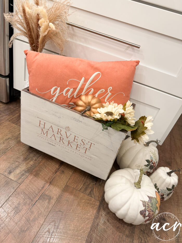 A white wooden Harvest Crate with Harvest Market text holds a burnt orange gather pillow, faux pumpkins, and flowers. White pumpkins are arranged beside it as charming Fall Decor, sitting on a wooden floor by white cabinets.
