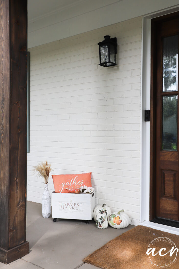 A front porch with a wood pillar, white brick wall, and a brown door. Fall decor includes a gather pillow, a Harvest Crate filled with flowers, white pumpkins, a glass vase with dried grasses, and a tan doormat.