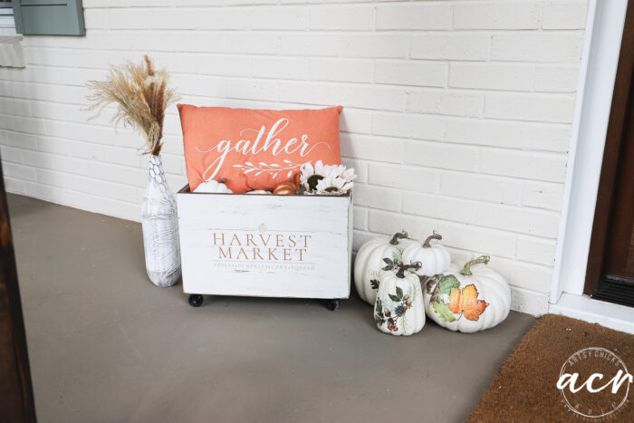 A fall-themed porch scene with white pumpkins, a vase of dried grass, and a gather pillow nestled in a Harvest Crate labeled Harvest Market, features charming Fall Decor completed by a round doormat on a gray floor beside a white brick wall.