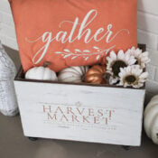 A white wooden Harvest Crate labeled Harvest Market holds white and gold pumpkins, white flowers, and an orange pillow with the word gather. Decorative vases and pumpkins add to the charming fall decor against a white brick wall.