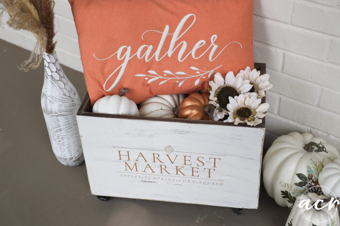 A white wooden Harvest Crate labeled Harvest Market holds white and gold pumpkins, white flowers, and an orange pillow with the word gather. Decorative vases and pumpkins add to the charming fall decor against a white brick wall.