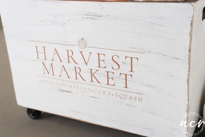 A white wooden Harvest Crate with “Harvest Market” in orange letters and the words apples, pumpkins, corn, squash below. The box has wheels and a small pumpkin graphic above the text—perfect as charming Fall Decor for your home.