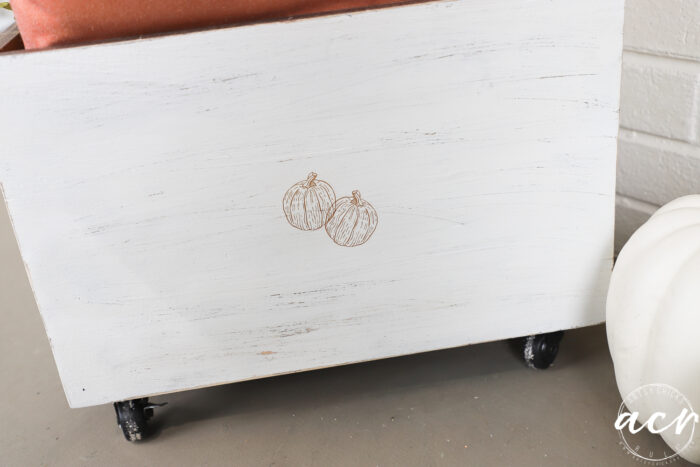 A white wooden planter box on wheels, styled as a charming Harvest Crate, features a simple sketch of two pumpkins on its front. A terracotta pot sits on top, and part of a white pumpkin is visible to the right—perfect for adding cozy Fall Decor to any space.