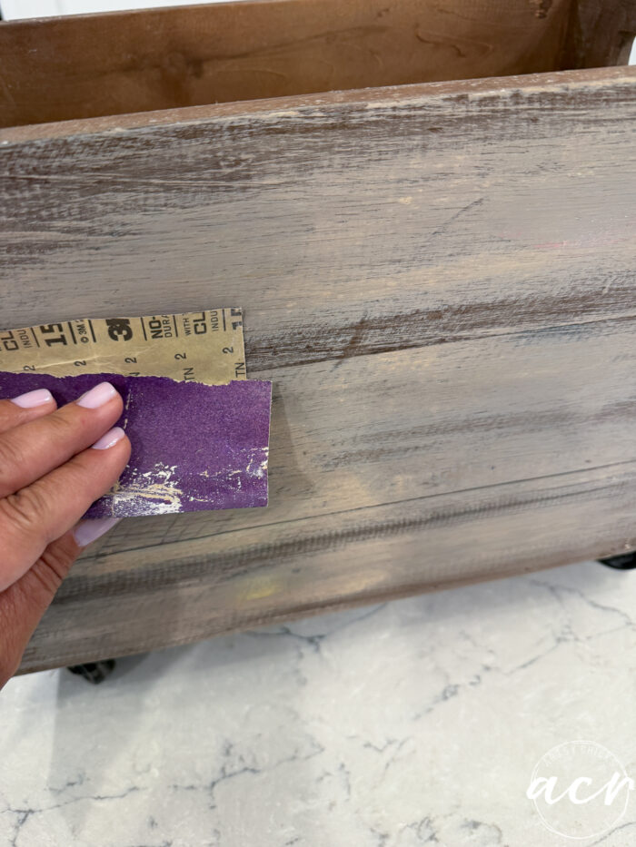 A hand holding sandpaper is sanding the side of a wooden drawer on a white marble surface. The weathered, painted finish of the drawer gives it the perfect rustic touch for creating a charming Harvest Crate, ideal for cozy Fall Decor.