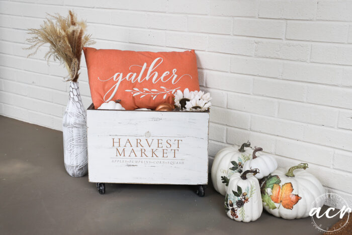 Autumn decor featuring a charming white Harvest Crate labeled “Harvest Market,” an inviting orange gather pillow, pumpkins, and a vase with dried wheat captures the essence of Fall Decor. White pumpkins adorned with delicate leaf designs are artfully arranged on a gray floor against a classic white brick wall, creating the perfect Fall Harvest display.