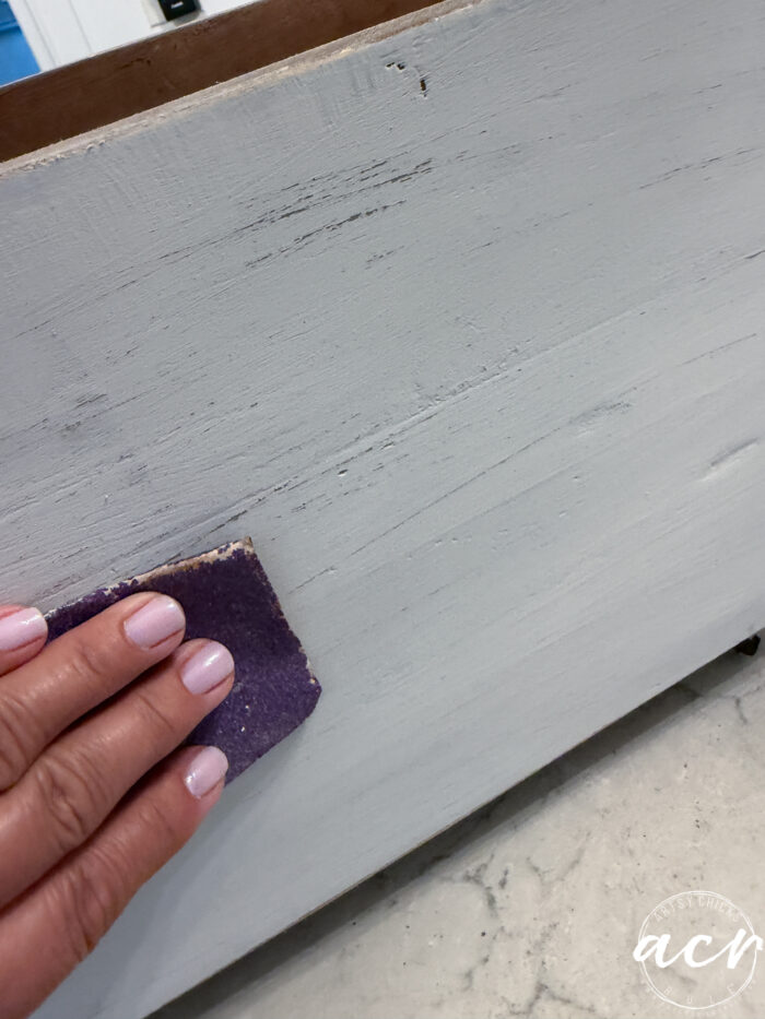 A hand with light pink nails sands a piece of painted wood using a small piece of sandpaper; the surface appears slightly distressed, making it perfect for transforming into Harvest Crate-inspired Fall Decor.