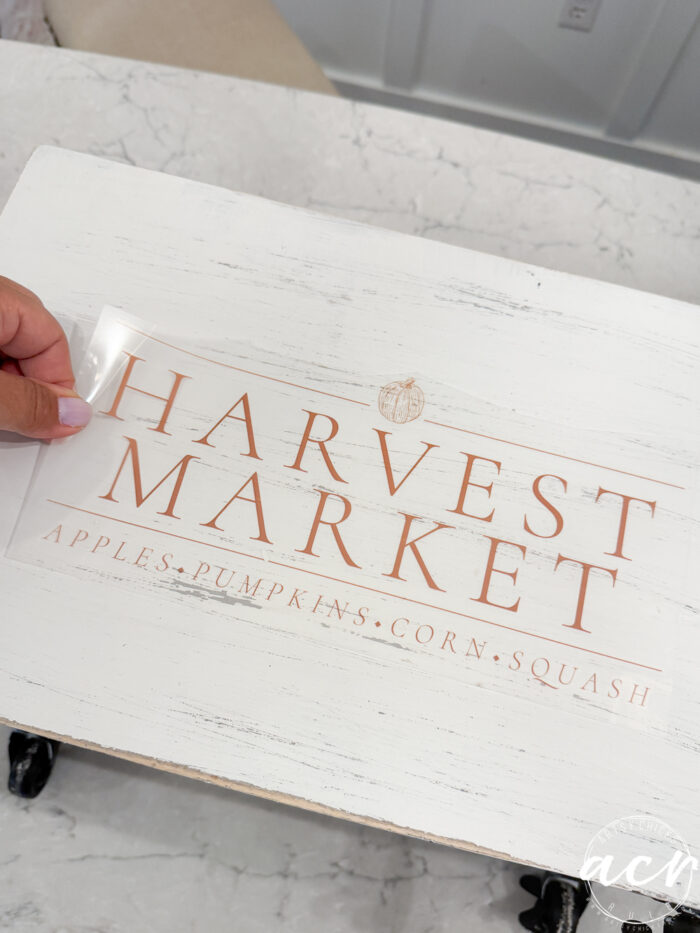 A hand places a clear decal reading HARVEST MARKET - APPLES • PUMPKINS • CORN • SQUASH on a white wooden surface, adding the perfect touch of fall decor to your harvest crate.