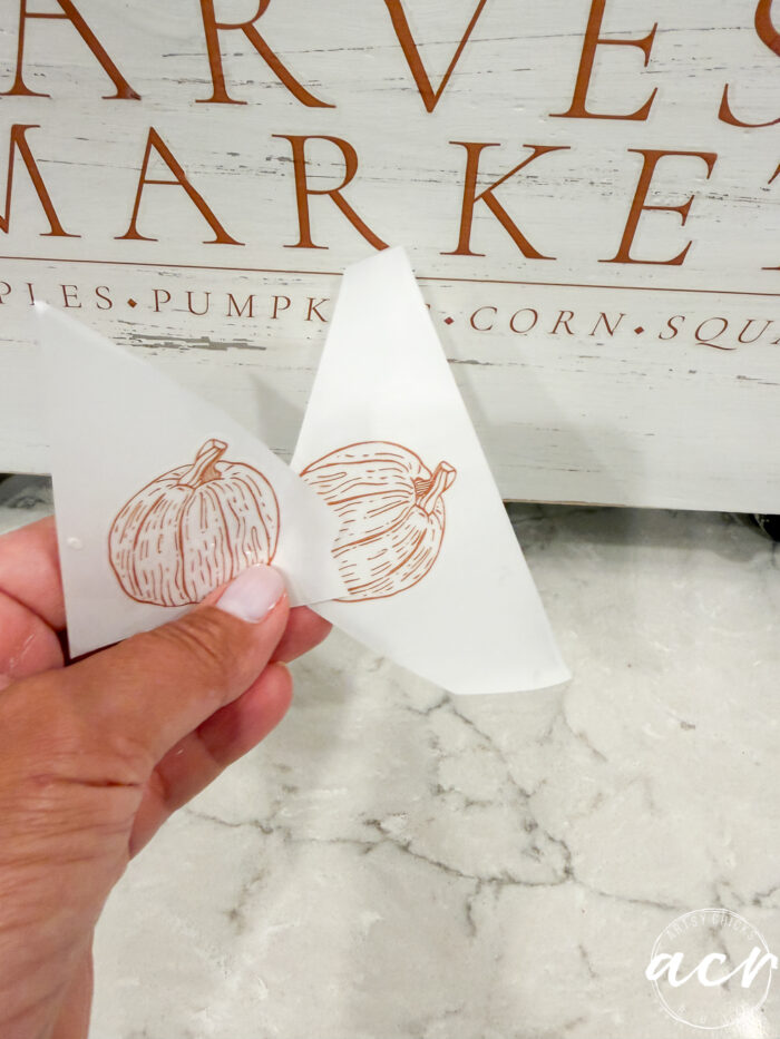 A hand holds a piece of transfer paper with a pumpkin design in front of a sign that reads HARVEST MARKET and lists various produce items, making it the perfect inspiration for cozy fall decor or creating your own harvest crate display.