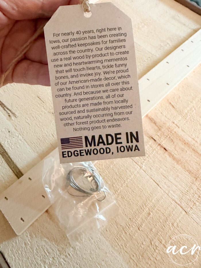 A hand holds a tag that reads about handcrafted keepsakes made in Edgewood, Iowa. Below, a bag with metal hardware rests on a stained wood countertop. The company emphasizes sustainability and American-made products.