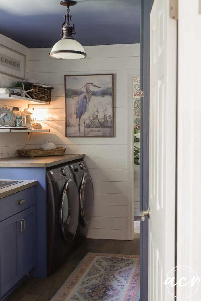 A laundry room with blue cabinets, a sleek wood countertop, two black front-load machines, white shiplap walls, a heron painting, shelves with decor, and a woven basket. A blue ceiling and patterned rug complete the coastal look.
