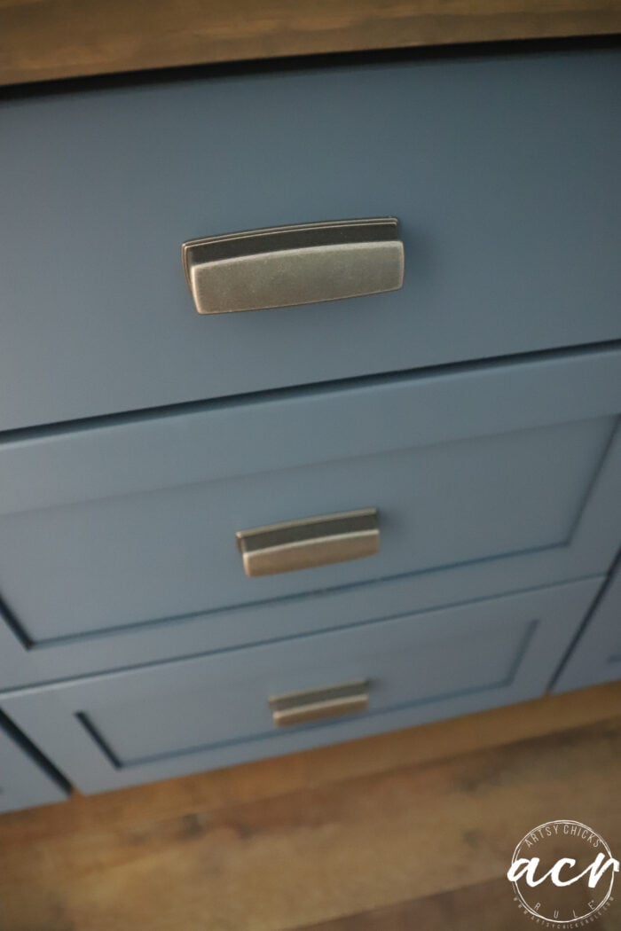 Close-up of three blue cabinet drawers with rectangular silver handles. The drawers are set against a wood countertop and wooden floor, showcasing a stylish countertop update perfect for any laundry room. A white circular logo is in the bottom right corner.