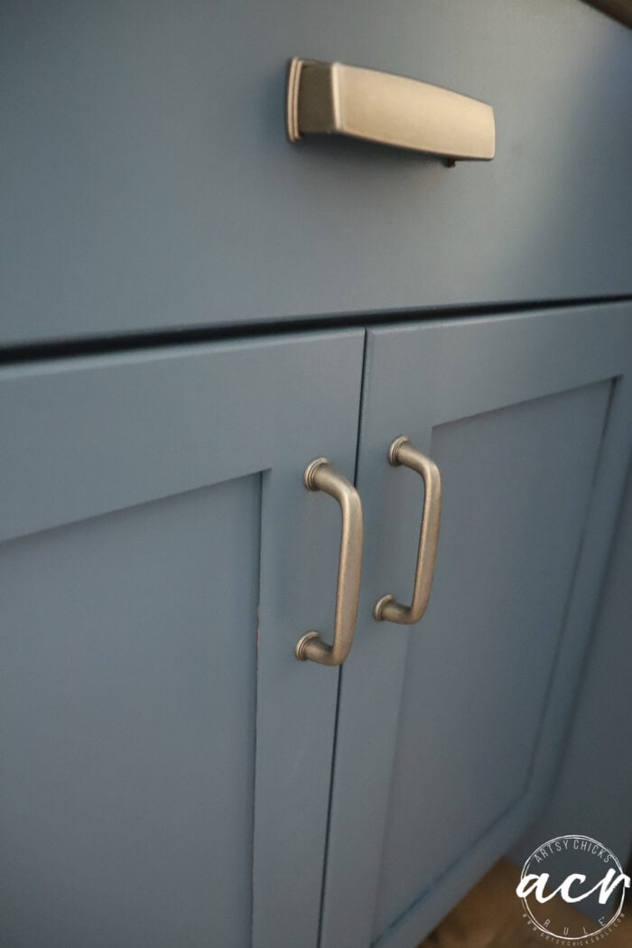 Close-up of blue cabinet doors with two silver handles and a silver drawer pull above, set beneath a sleek wood countertop. A white logo with ACR is in the bottom right corner.