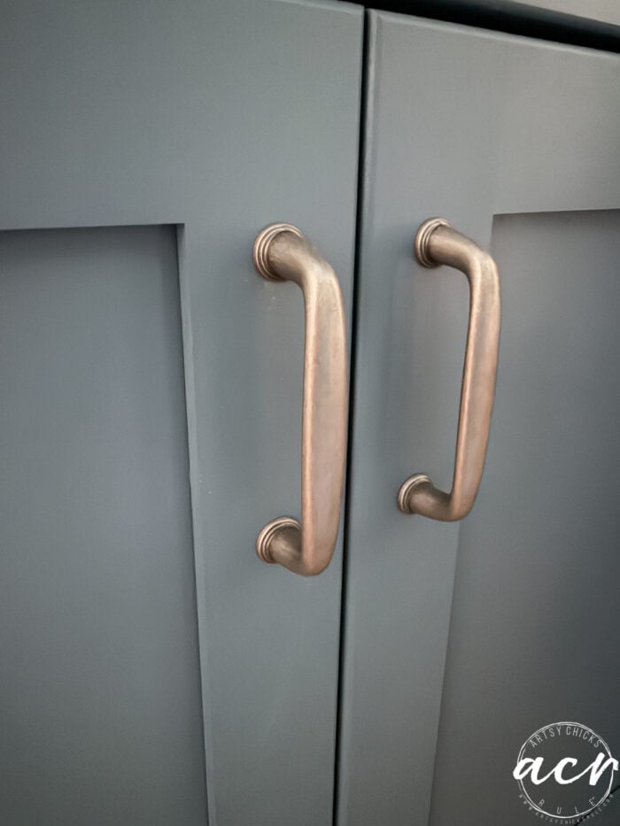 Close-up of two copper-colored cabinet handles on matte blue-gray cabinet doors, perfect for a laundry room makeover or a countertop update. A small circular logo with cursive text is visible in the lower right corner.