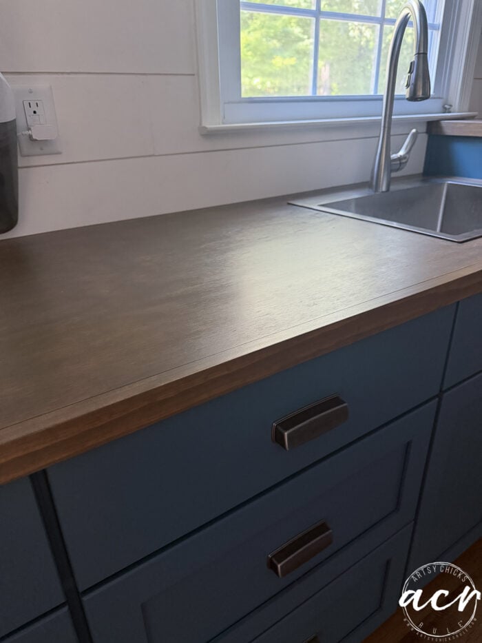 A kitchen countertop with a wood countertop surface, blue drawers, a modern faucet, and a window above the sink letting in natural light offers an inviting space for meal prep or even a stylish countertop update.