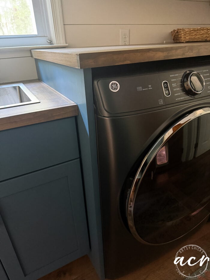 Front-loading black washing machine next to blue cabinetry with a wood countertop, placed beneath a window in a laundry room.