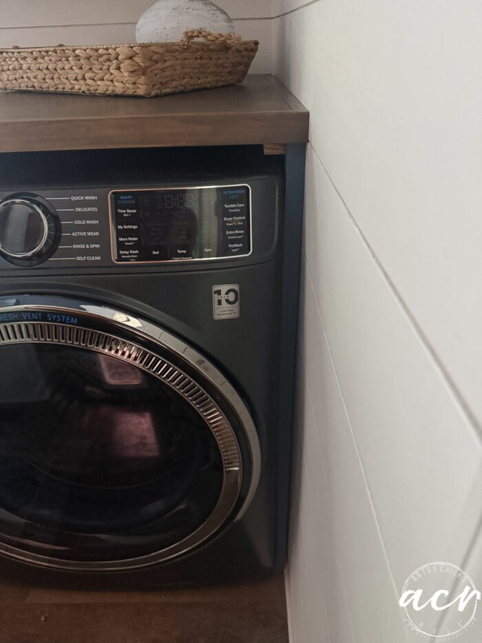 A close-up of a dark-colored front-loading washing machine with a digital display and cycle options in a stylish laundry room, positioned next to a white shiplap wall and topped with a woven basket sitting on a warm wood countertop.