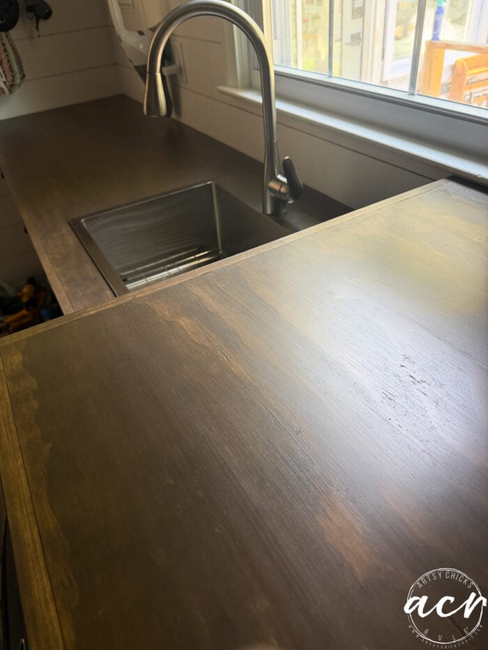 A close-up of a wood countertop with a sleek finish next to a metal sink and a modern faucet, under a window with sunlight streaming through. Perfect for a laundry room refresh or countertop update, this space combines style and practicality. A small circular ACR watermark is in the bottom right corner.