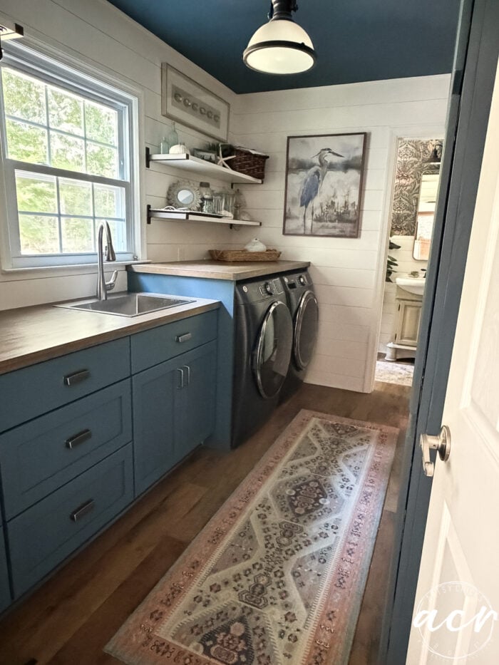 Laundry room with blue cabinets, a recent wood countertop update, stainless steel washer and dryer, open shelves with decor, a window, patterned rug, and bird artwork on white shiplap walls. Door leads to another room.