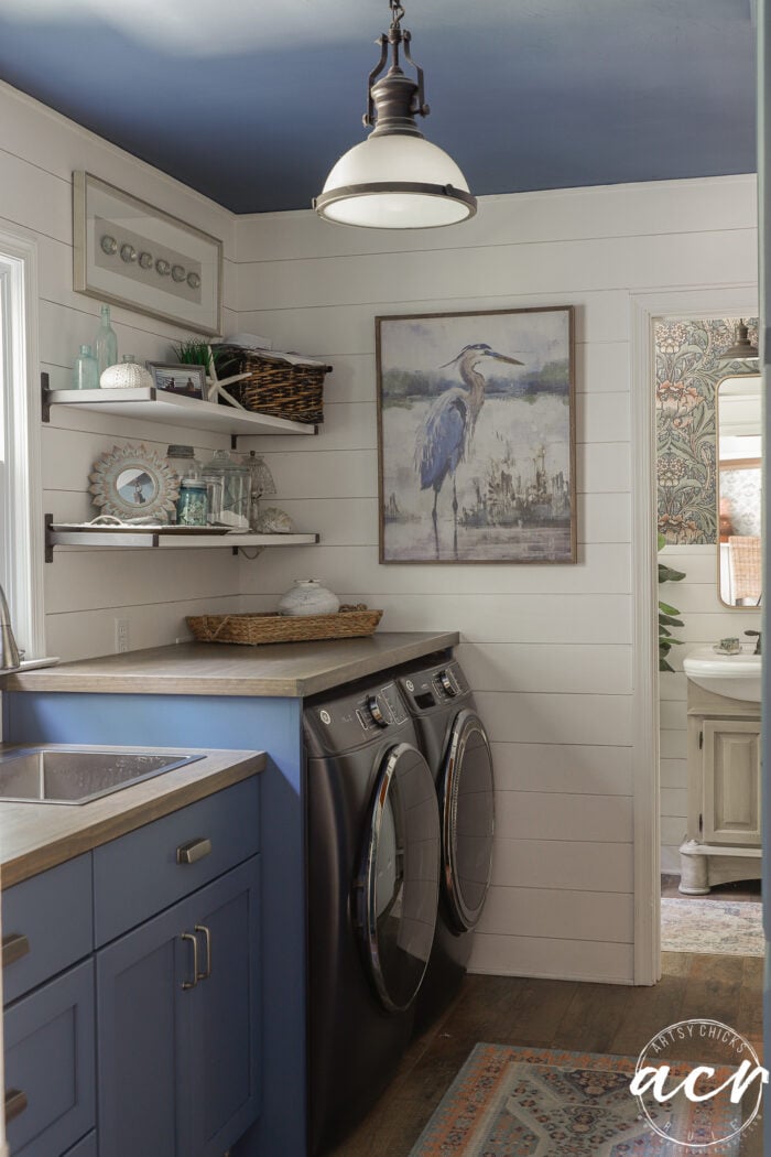 Laundry room with blue cabinets, a sleek wood countertop, black washer and dryer, open shelves with decor, a heron painting on the wall, and a rustic rug on wooden floor. A bathroom is visible through an open door.
