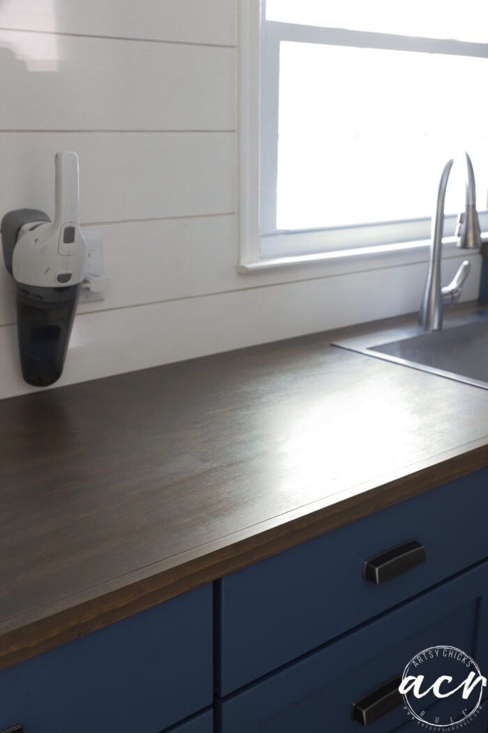 A clean wood countertop with blue cabinets, a stainless steel sink, modern faucet, wall-mounted handheld vacuum, and a window above the sink letting in natural light—perfect for an updated laundry room.