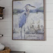 Laundry Room Update #2 (stained wood countertop, art and hardware) Framed painting of a blue heron standing in shallow water with greenery in the background, hung on a white shiplap wall above a laundry room featuring a stylish wood countertop.