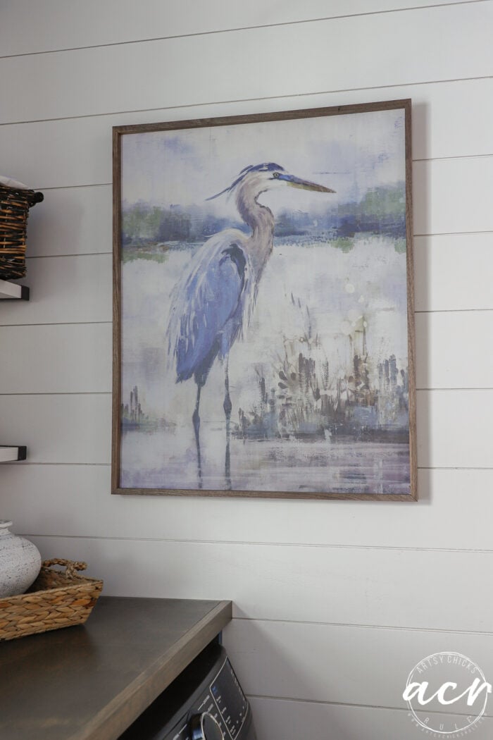 Framed painting of a blue heron standing in shallow water with greenery in the background, hung on a white shiplap wall above a laundry room featuring a stylish wood countertop.