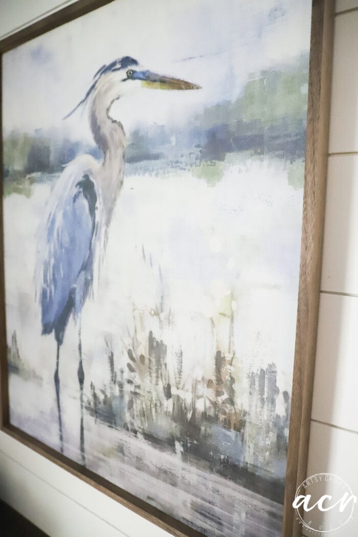 Painting of a blue heron standing by water, surrounded by a soft green and white background, framed with wood—a serene piece perfect to update your laundry room, especially when paired with a warm wood countertop.