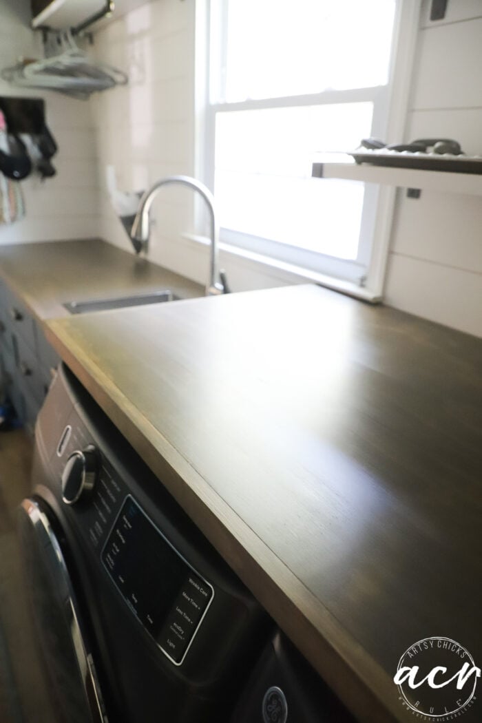 Enjoy a stylish countertop update with this wood countertop installed over a front-load washing machine in a laundry room, complete with a sink, shelves, and window in the background.
