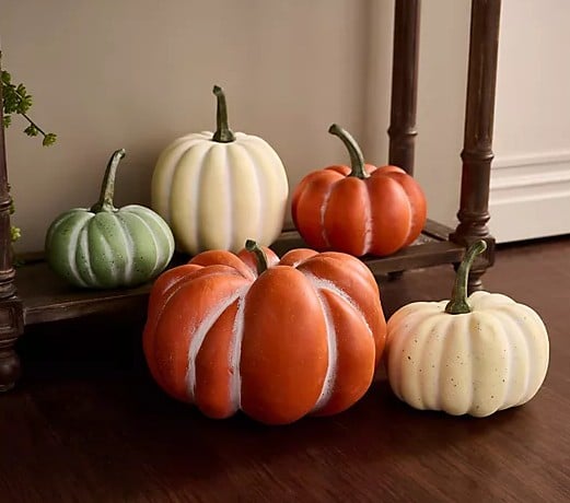 Five decorative pumpkins in orange, white, and green—true Fall Favorites—are arranged on a wooden floor and a small step next to a banister, creating an autumn-themed display.