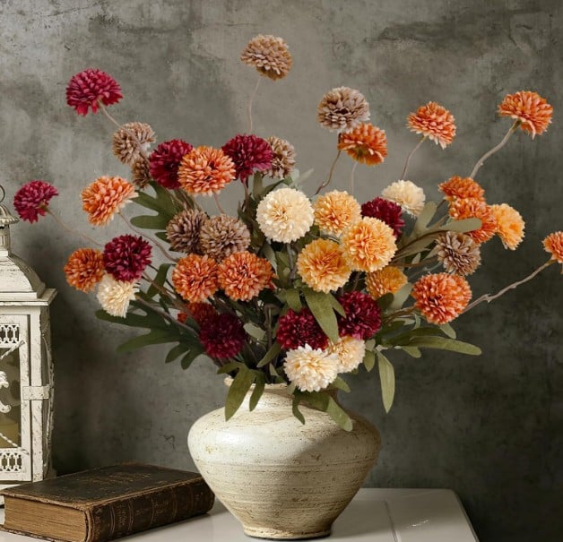 A beige vase holds a bouquet of artificial pompom flowers in shades of red, orange, cream, and brown—perfect fall favorites. The vase sits on a white table beside a vintage lantern and a closed book, against a gray textured wall.