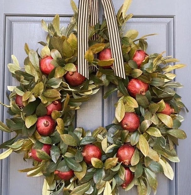 A wreath made of green leaves and red pomegranates hangs on a gray door with a striped ribbon at the top, capturing the Best of Fall with its rich, seasonal charm.