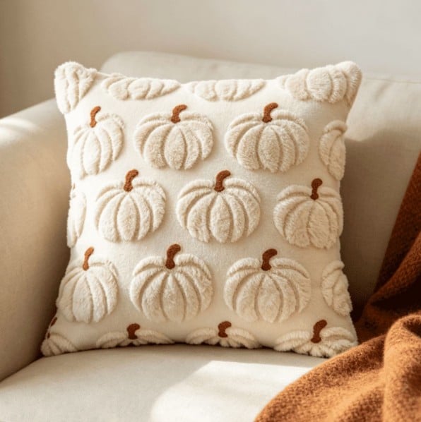 A cream-colored pillow with raised, textured pumpkin designs and brown stems—one of our fall favorites—sits on a light beige couch next to a rust-colored blanket.