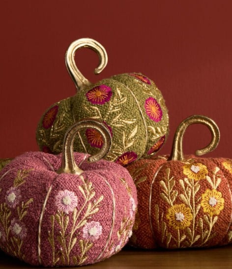 Three decorative fabric pumpkins with embroidered floral designs in pink, green, and orange sit against a red background. Each pumpkin has a gold stem and detailed stitching—perfect for showcasing your Fall Favorites in any cozy setting.