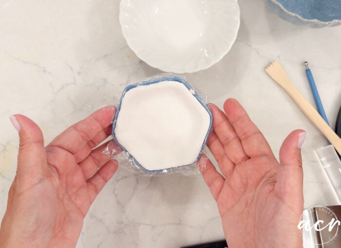 Two hands hold a hexagonal blue dish lined with plastic wrap, containing white air dry clay. A white bowl and pottery tools are on a white marble surface in the background, perfect for comparing brands or experimenting with different clay brands.