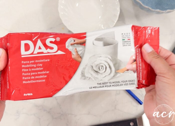 Hands hold a red and white package of DAS Air Dry Clay, showing images of a hand shaping clay and finished clay objects on the package. The surface underneath is a light, veined countertop—a great scene for anyone interested in clay brands comparison or searching for the best air dry clay.