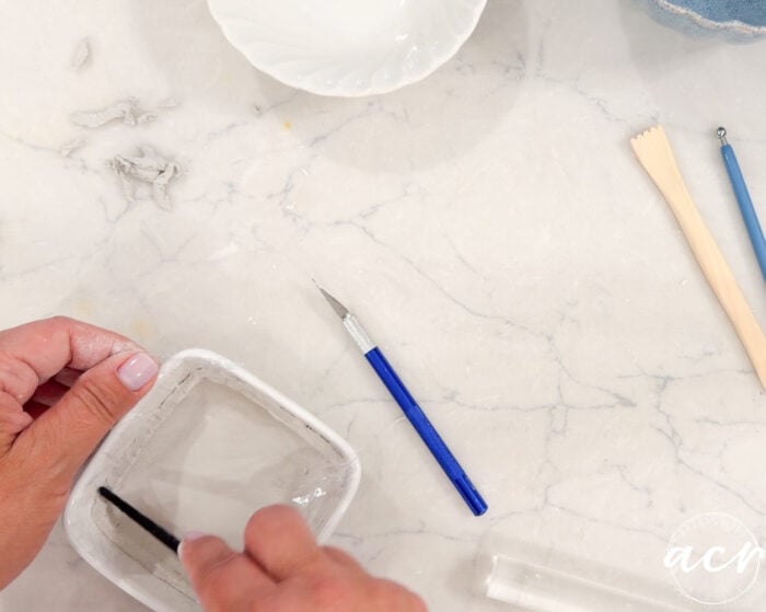 A person smooths air dry clay in a small white dish with a tool. Sculpting tools and a ceramic bowl are nearby on a marble surface.