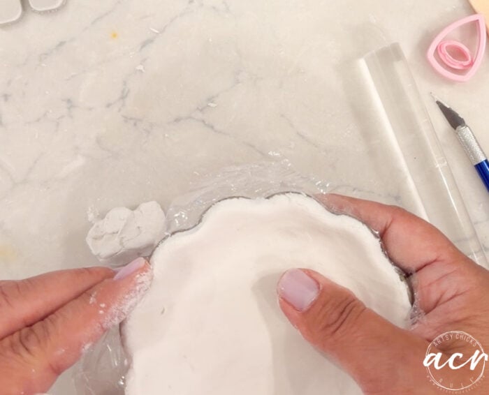 Hands shaping white air dry clay on a scalloped-edge surface, with sculpting tools and plastic wrap nearby, on a light marble countertop—perfect for anyone interested in comparing brands or reading a detailed clay review.