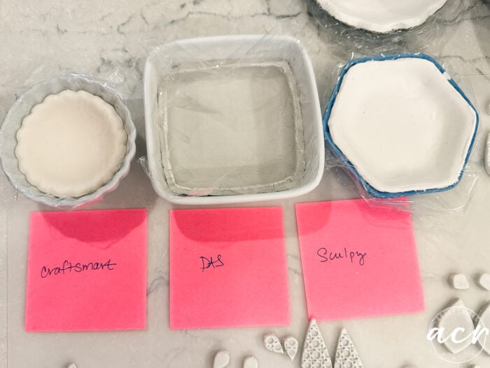 Three white ceramic dishes contain clay in different shapes, each labeled with a pink sticky note: Craftsmart, DAS, and Sculpey. Small clay pieces are nearby on a marble surface, providing a clear visual for this Air Dry Clay Review and Clay Brands Comparison.