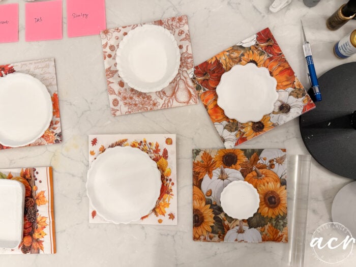 Four square coasters with autumn-themed designs, each holding a round white dish, are on a marble surface. Nearby, craft tools, a roller, and pink sticky notes suggest an ongoing project—perfect for experimenting with different Air Dry Clay brands in your next creative session.