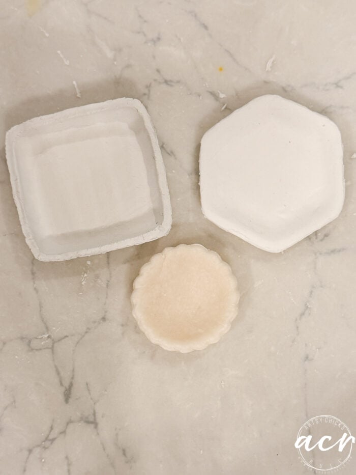 Three small, unfinished air dry clay dishes—one square, one hexagonal, and one round—sit on a light marbled surface. The round dish appears more textured than the others, showcasing the unique qualities often found in different air dry clay brands. A watermark ACR is in the bottom right corner.